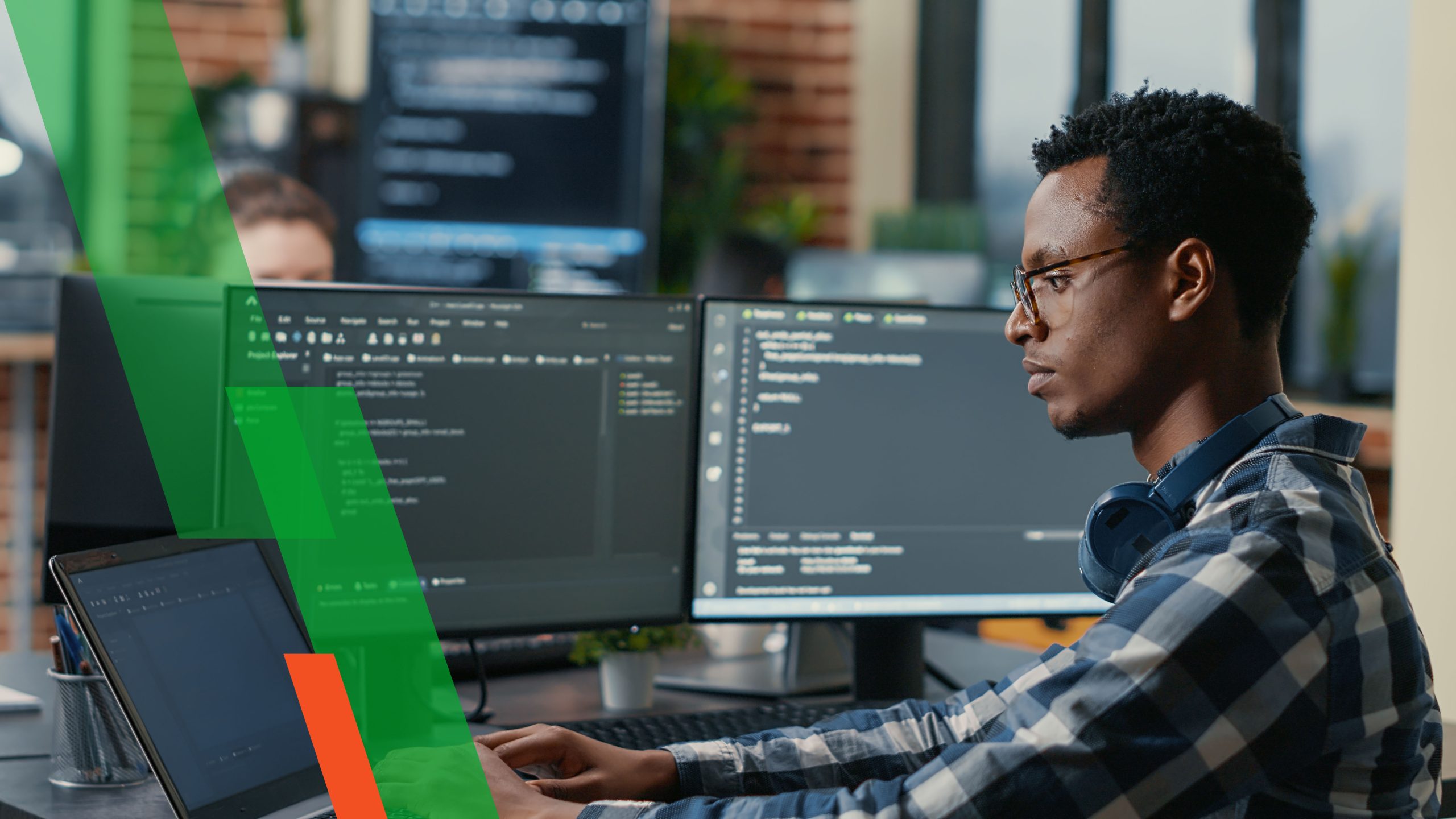 Sofware developer thinking while touching beard while typing on laptop sitting at desk with multiple screens parsing code. Focused database admin working with team coding in the background.